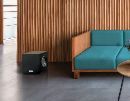Zwarte kubusvormige subwoofer op grijze vloer, blauwe bank met houten frame, houten muur op de achtergrond, gedeeltelijke ronde tafel