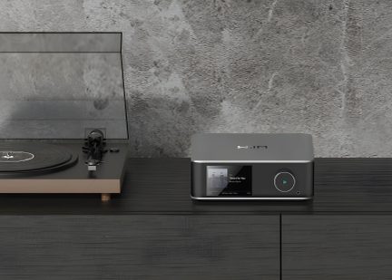 Gramofon s prozornim pokrovom na levi, digitalni avdio predvajalnik z zaslonom in okroglim gumbom na desni, temna lesena površina