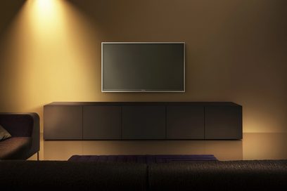 Lavt, mørkegråt TV-møbel med stort, rektangulært, fladt sort TV-skærm i et oplyst rum med orange-gule vægge