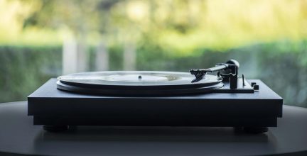 Sort pladespiller med tonearm og gennemsigtig vinylplade, set fra siden, sløret baggrund med vegetation og naturligt lys.