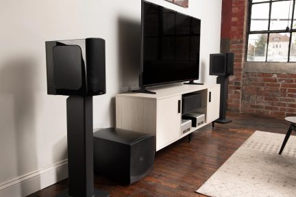 Soggiorno moderno con home theater, TV, altoparlanti, divano nero, muro di mattoni, pavimento in legno, tappeto beige