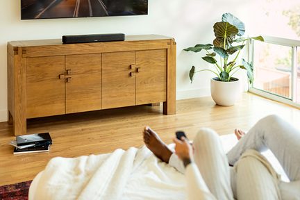 Black Denon soundbar on a light wood TV stand in a bright living room, with a green plant and a wall-mounted television
