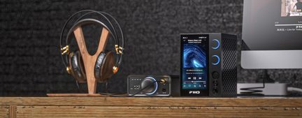 Audio set featuring black over-ear headphones with gold accents resting on a wooden stand, a small rectangular black and silver DAC amplifier with a connected knob, and a black amplifier player with a color display, dual blue-ringed knobs, and a perforated front panel, front view on a wooden desk