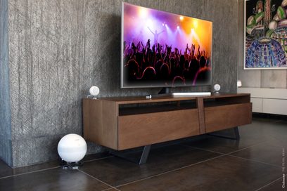Koka TV skapītis ar televizoru, kurā redzama koncertā esošā ļaužu pūlis krāsainā fonā. Uz grīdas divas baltas sfēriskas lampas