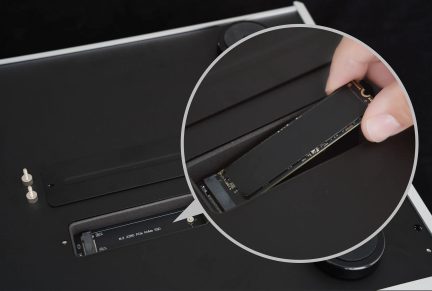 Matinės juodos spalvos aliuminio elektroninis korpusas, vaizdas iš viršaus, nuimtas prieigos skydelis atidengia M.2 2280 PCIe NVMe SSD lizdą su dviem sidabriniais kniedėtais varžtais ir apvaliu juodu guminiu kojeliu