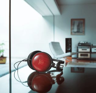 Kapalı, kulak çevreleyen, ahşap kaplamalı Audio Technica ATH-AWAS kulaklık, modern bir iç mekânda cam bir masa üzerinde duruyor.