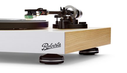 Giradischi con base rettangolare in legno chiaro, frontale bianco e logo "Roberts", ripreso in controplongée con visuale obliqua che mostra il piatto circolare nero, il braccio di lettura nero con finitura carbonio e testina verde, e i piedi regolabili neri