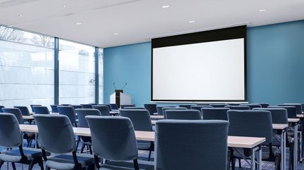 Konferencelokale med rækker af blå stole vendt mod en stor hvid projektionsskærm på en lyseblå væg