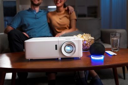 Optoma UHZ50 hvid projektor stillet på et træbord, smilende par sidder på en grå sofa i baggrunden