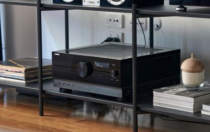 Čierny Yamaha zosilňovač domáceho kina s veľkým ovládačom hlasitosti a predným displejom na modernom audio regáli