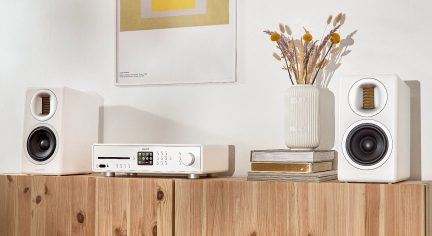 Cadena de música blanca con reproductor de CD, amplificador y altavoces, colocada sobre un mueble de madera clara, con un jarrón de flores secas