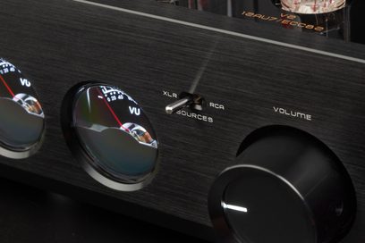 Primer plano, vista frontal en ligera picada de la fachada de aluminio cepillado negro del preamplificador de auriculares de alta fidelidad Cayin HA-3A, con dos vúmetros abombados, un selector de palanca XLR/RCA central y un gran botón de volumen a la derecha