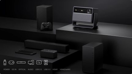 Sort og grå projektor på drejeligt beslag, omgivet af spillekonsoller, en soundbar og en computer