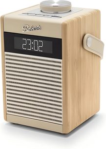 Rádio Roberts Rambler Midi v krémové barvě, krychlový dřevěný pouzdro v světlém tónu s krémovými plastovými panely a boční rukojetí, pohled z mírně zvýšeného tříčtvrtečního úhlu, přední strana zdobená perforovanou krémovou mřížkou, černý digitální displej zobrazující čas a velký stříbrný kruhový knoflík na horní straně