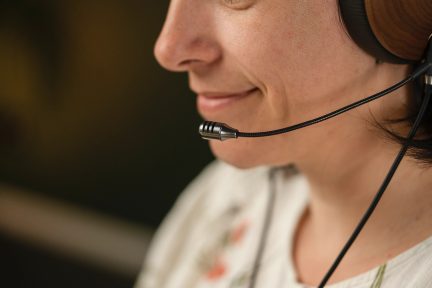 Sivuprofiili, osittain näkyvät kasvot, ruskeat kuulokkeet, musta joustava mikrofoni, valkoinen paita kukkakuviolla, sumea tausta