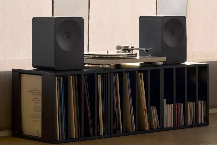 Vinilinė plokštelių grotuvas su dviem juodomis kolonėlėmis ant juodos plokštelių spintelės, vaizdas iš priekio moderniame salone