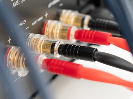 Detailní šikmý záběr na řadu konektorů reproduktorů z průhledného plastu a pozlaceného kovu na audio panelu, vybavených červenými a černými banánkovými konektory