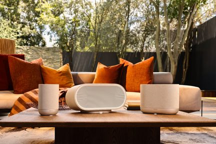 Beige trådløse Denon-højttalere på et lavt sofabord i træ foran udendørs sofa med orange puder