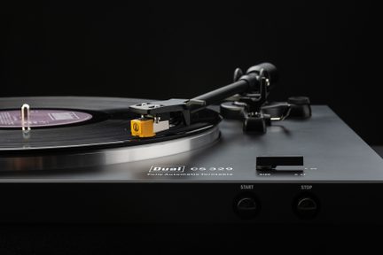 Platine tourne-disque noire mate en métal et plastique, plateau circulaire argenté supportant un vinyle noir, bras de lecture noir terminé par une cellule jaune, vue rapprochée frontale légèrement plongeante
