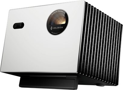 Valerion StreamMaster Plus 2 projector, matte white rectangular metal casing with a black perforated side grille and a circular lens with a black ring on the front, shown in a front-right perspective with a slight low-angle view
