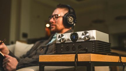 Ampli casque hi-fi argenté avec écran central et deux gros boutons, posé sur table, audiophile au casque en arrière-plan