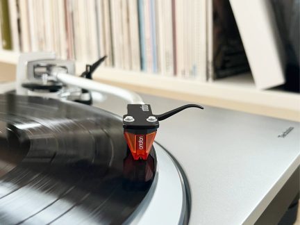 Cellule phono Ortofon noire et orange en plastique et métal sur disque vinyle noir, bras de platine argent, gros plan