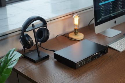 Schwarzer Focal Kopfhörer auf Metallständer, verbunden mit schwarzem Questyle Kopfhörerverstärker auf Holzschreibtisch mit iMac