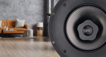 Close-up of a circular black speaker on the right, orange sofa, white cushions, coffee table, lamp, and wooden floor in a blurred background