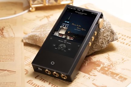 Lettore audio digitale rettangolare in metallo nero opaco, vista di tre quarti anteriore destra, schermo a colori a tutta faccia, rotella del volume zigrinata dorata in alto sul lato destro, tre pulsanti dorati sotto di essa e tre prese jack più una porta USB sul bordo inferiore