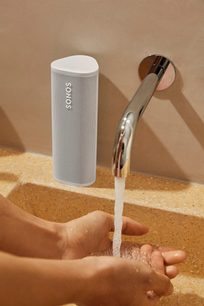 Bílý bluetooth reproduktor Sonos Roam SL, otevřený vodovodní kohoutek nad rukou držící bílý válcový reproduktor