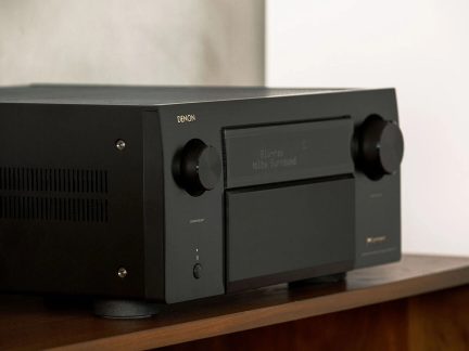 Černý kovový audio-videový zesilovač, pohled z levého předního tříčtvrtečního úhlu, se dvěma velkými symetrickými otočnými knoflíky, centrálním displejem a bočními větracími otvory, umístěný na dřevěné polici