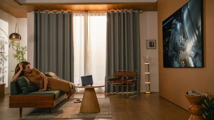 Stor väggmonterad TV i modernt vardagsrum, kvinna liggande på grön soffa, grå gardiner och varm inredning i trä
