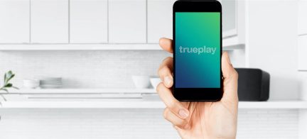 Smarttelefon som viser logoen "trueplay" holdt i en hånd mot en hvit kjøkkeninnredning i bakgrunnen
