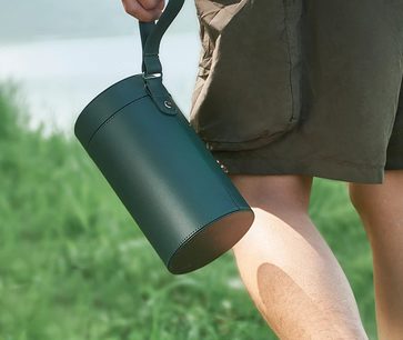 Cilindro verde scuro con manico, tenuto da una mano, sfondo di erba verde, gamba in pantaloncini marroni visibile