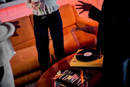 Svetlá drevená gramofónová platňa s transparentným krytom a čiernou vinylovou platňou na okrúhlom drevenom stole, v pozadí oranžová pohovka