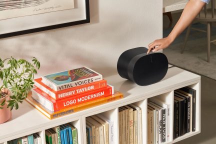Oval black speaker placed on a white shelf next to a stack of colorful books, a woman's hand adjusting the speaker