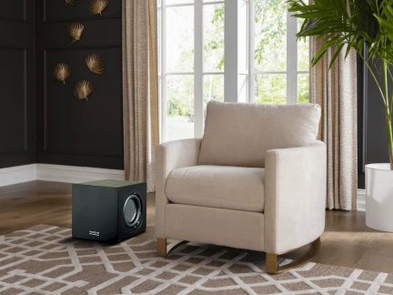 Beige fauteuil met gewatteerde armleuningen, zwarte subwoofer, zwart versierde muur, beige gordijnen, groene plant, tapijt met geometrische patronen