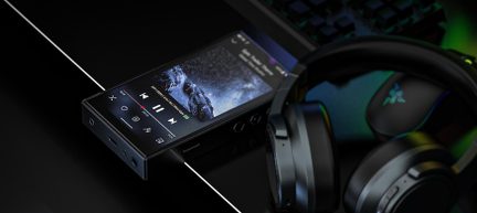 Sort digital musikafspiller i metal med tændt touchskærm og synlige sekskantede sideknapper, eksponerede porte i bunden, placeret på skrå på et sort skrivebord med sorte hovedtelefoner delvist i forgrunden, set fra en skrå vinkel