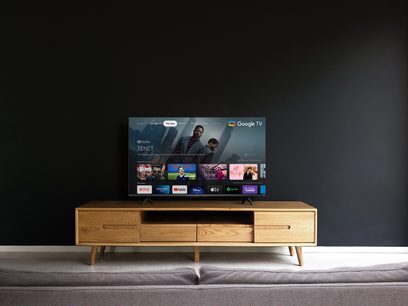TCL 43QLED760 2022 fladskærms-TV på lyst trækabinet, skærmen viser Google TV-grænsefladen med miniaturebilleder af film og tv-serier