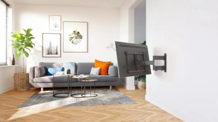 Svart väggfäste för TV med ledad arm, TV sedd bakifrån och riktad mot en grå soffa i ett ljust vardagsrum
