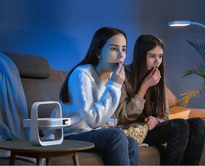 Twee personen zitten op een bank, eten popcorn, staande lamp aan, witte projector op salontafel