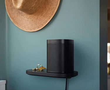 Czarna głośnik bezprzewodowy Sonos One na czarnym uchwycie ściennym Sonos Shelf. Słomkowy kapelusz zawieszony na jasnej niebieskiej ścianie