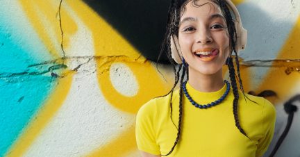 Osoba zepředu, sluchátka béžová, žluté tričko, náhrdelník z modrých perel, pozadí zeď s graffiti v žluté a modré barvě