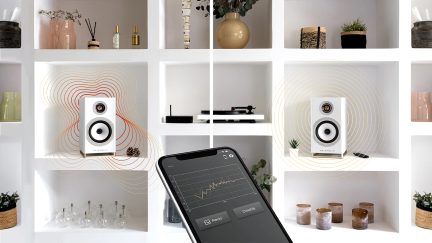 Rafturi albe cu difuzoare albe, obiecte decorative, smartphone afișând un grafic, pick-up negru pentru viniluri