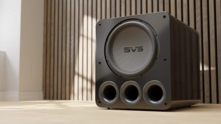 Bassreflexový reprobox v matné černé barvě s centrálním kruhovým mřížkovým krytem a 3 kulatými výstupními otvory na přední straně, perspektivní pohled na světlém podkladu