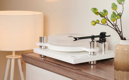 Biely lakovaný vinylový gramofón s priehľadným akrylovým tanierom, kovovými valcovými nohami a čiernym ramienkom, pohľad z troch štvrtín z mierneho nadhľadu