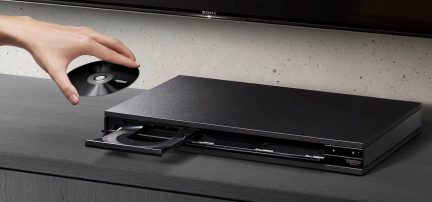 Sort Blu-ray-afspiller med åben skuffe, sort disk holdt af en hånd, flad overflade, grå væg i baggrunden