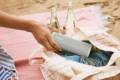 Sonos Roam Lunar White speaker placed on a red and white striped beach towel, next to two glass bottles