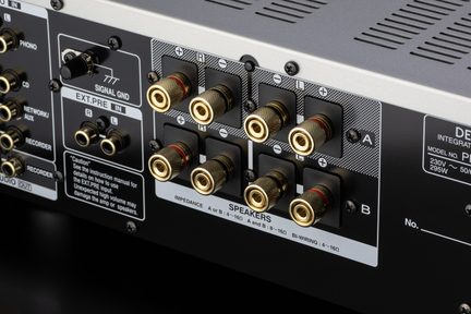 Zadní pohled zblízka na černý a stříbrný kovový audio zesilovač, osm pozlacených rýhovaných svorkovnic pro reproduktory s červenými a černými kroužky uspořádaných do dvou řad