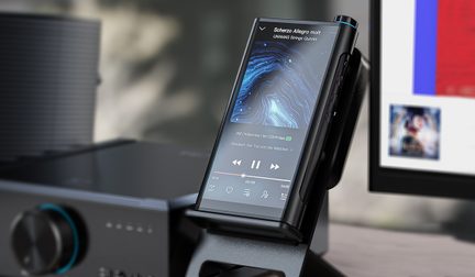 Sort, metallisk bærbar afspiller set i trekvart vinkel forfra, tændt touchskærm med musikinterface, tre synlige runde sideknapper og øverste drejehjul, placeret på en plastikholder
